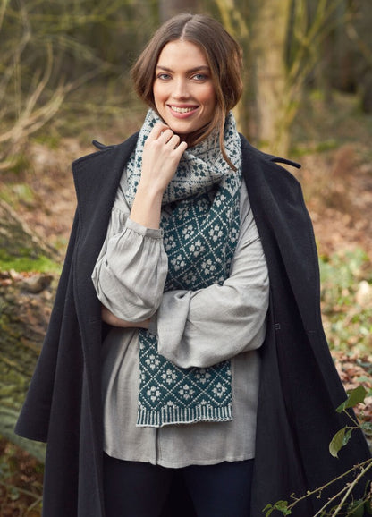 Woman smiles in a forest wearing a patterned scarf, light gray blouse, and dark coat. The scene feels cozy and serene, with soft autumn colors.