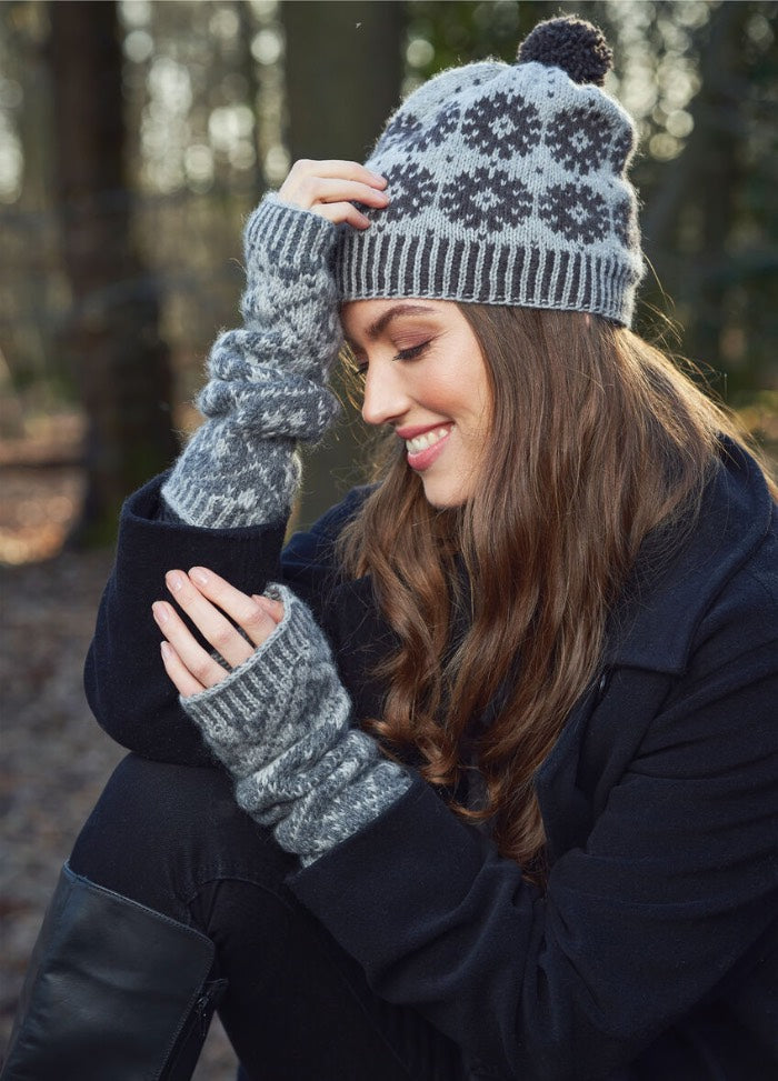 A woman in a patterned winter hat and matching fingerless gloves smiles and sits outdoors. The setting is a serene, sunlit forest.