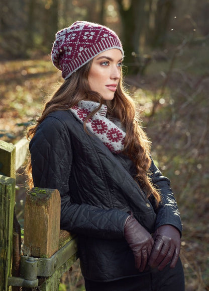 Young woman in a forest, wearing a maroon knit hat and scarf with white snowflake patterns, leaning on a wooden fence. She looks thoughtful.