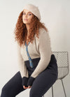 A woman with long curly auburn hair wears a cropped cardigan with beige upper body, dark navy ribbed cuffs and hem, and a denim blue button band. She accessorises with a soft beige beret and sits on a modern wire-frame chair against a plain background.