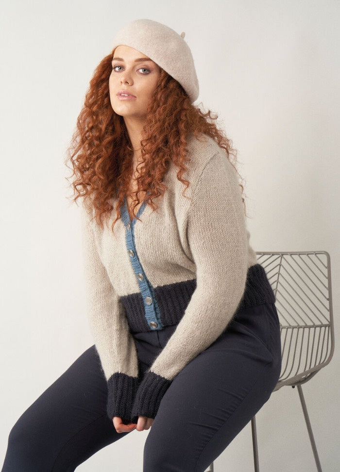 A woman with long curly auburn hair wears a cropped cardigan with beige upper body, dark navy ribbed cuffs and hem, and a denim blue button band. She accessorises with a soft beige beret and sits on a modern wire-frame chair against a plain background.