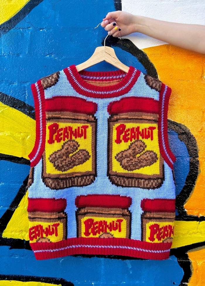 Knitted vest with a colorful peanut butter jar design hangs against a bright blue and yellow mural. A hand holds the hanger. Playful and vibrant vibe.