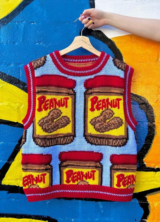 Knitted vest with a colorful peanut butter jar design hangs against a bright blue and yellow mural. A hand holds the hanger. Playful and vibrant vibe.