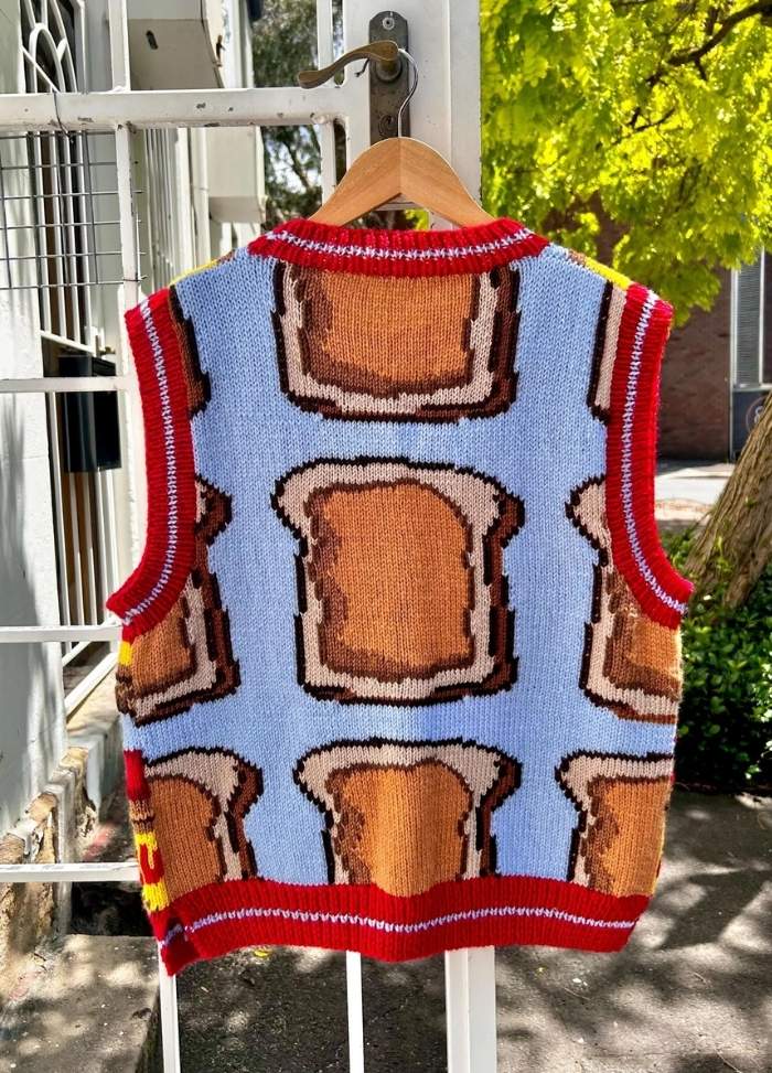 A knitted vest with a playful pattern of toast slices on a light blue background, bordered with red trims. It hangs outside on a wooden hanger.