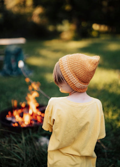 Orange knit beanie worn by a child near a campfire, with green grass and trees in the background.