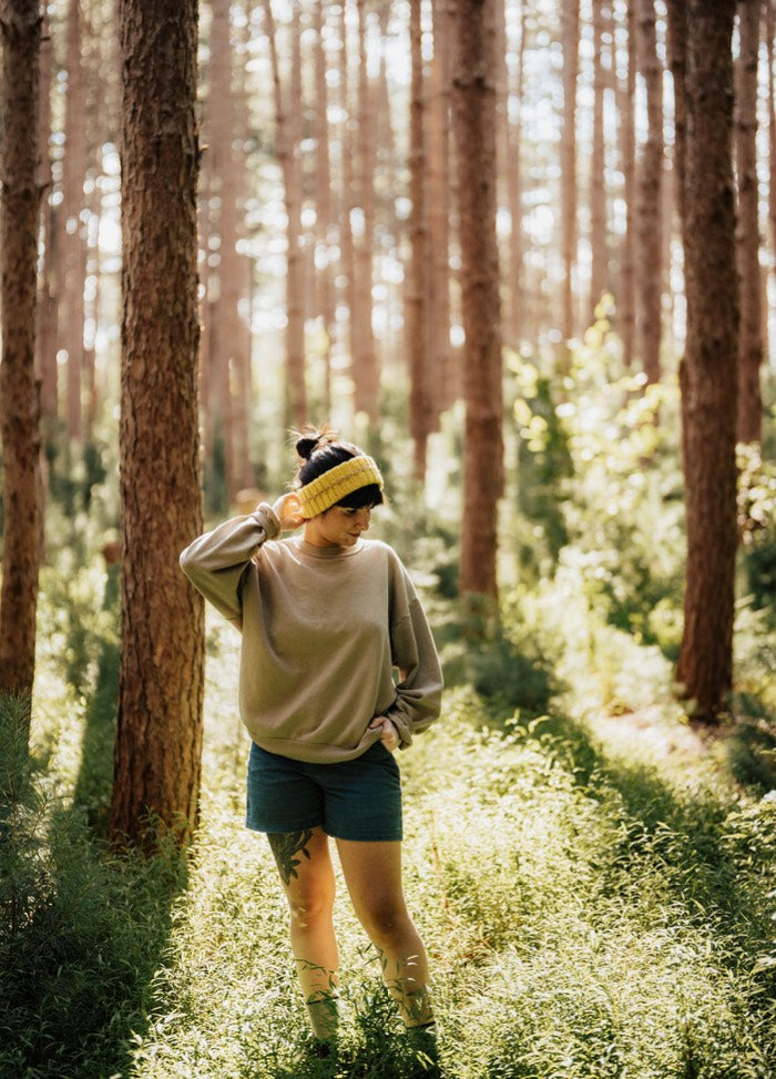 Yellow knit headband worn in a sunlit forest with a beige sweater and blue shorts.