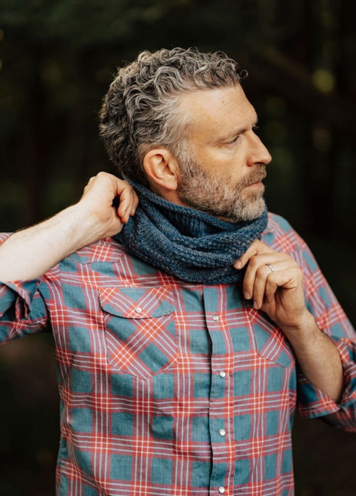 Blue knitted cowl worn with a red and blue plaid shirt in a forest setting.