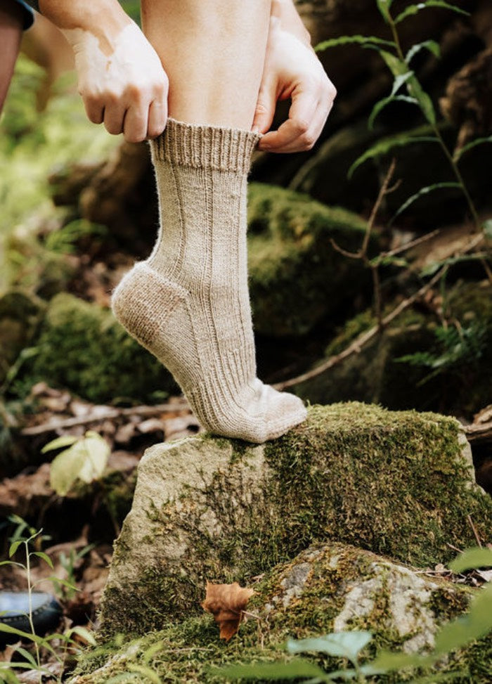 Thick beige woolen socks being pulled up on a mossy rock in a forest setting.