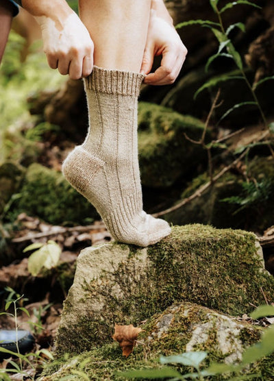 Thick beige woolen socks being pulled up on a mossy rock in a forest setting.