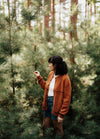 Orange-brown knitted jacket worn in a pine forest, paired with a white shirt and denim shorts.