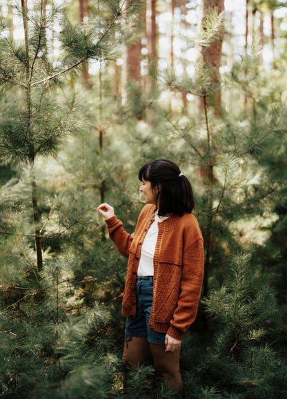 Orange-brown knitted jacket worn in a pine forest, paired with a white shirt and denim shorts.