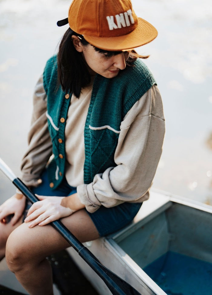 Green buttoned vest worn in a boat, paired with a beige shirt, blue shorts, and orange ‚ÄúKNIT‚Äù cap.