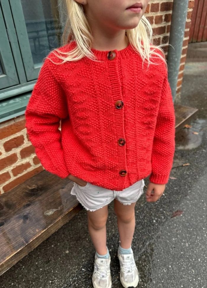 A young girl stands outdoors wearing a vibrant red knit sweater with wooden buttons, white shorts, and sneakers. The brick wall behind her adds texture.