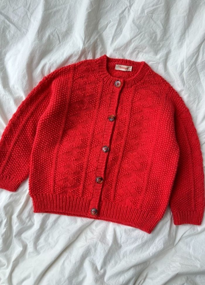 A vibrant red knitted cardigan with a textured pattern, featuring brown buttons, lies on a white fabric background, evoking a cozy and warm feel.