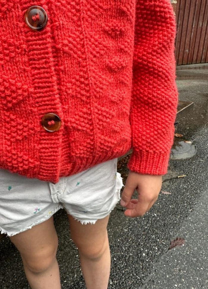 Child wearing a textured red knit sweater with large buttons and light denim shorts, standing on a wet asphalt surface. The mood is casual and cozy.