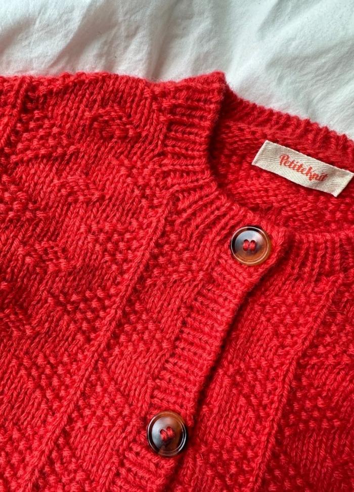 Red knitted sweater on a white background, featuring a detailed pattern, round neckline, and two dark buttons. A small label is sewn near the collar.