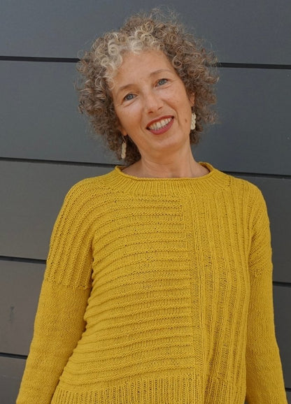 Smiling woman with curly hair wearing a textured mustard yellow sweater. She stands against a dark gray panel wall, conveying a joyful and warm tone.