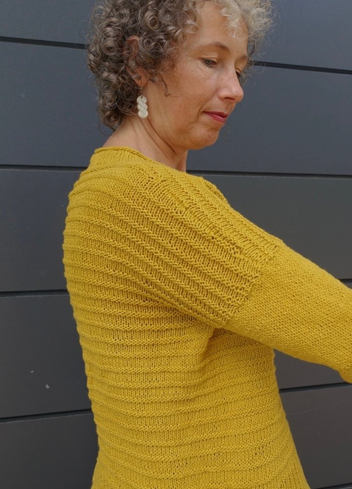 A person with curly hair wears a textured, mustard-yellow knit sweater standing against a dark gray paneled wall, conveying a calm, reflective mood.