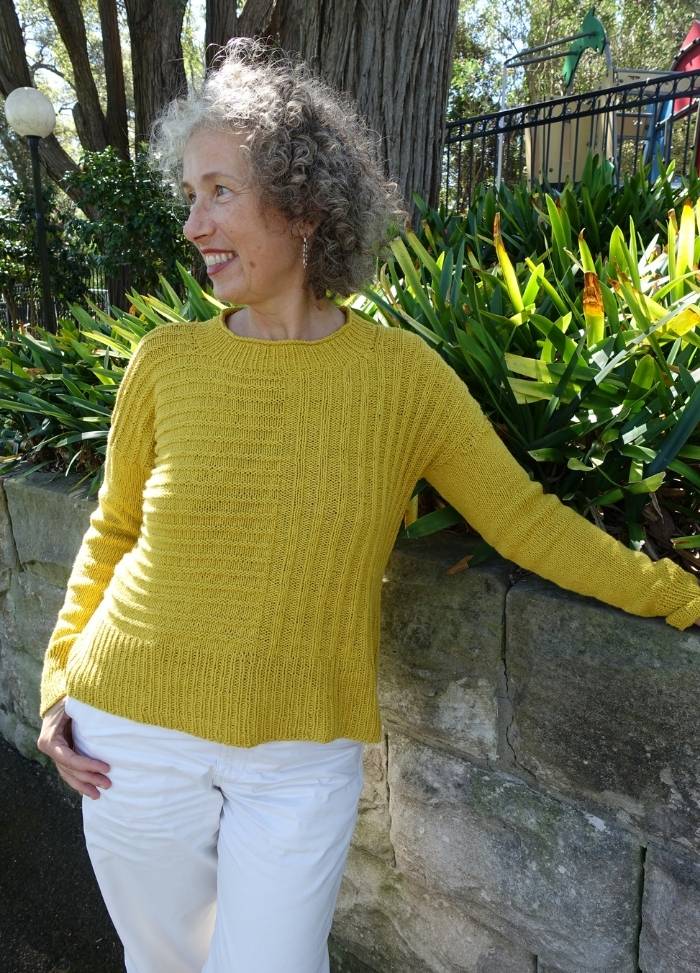 A woman with curly gray hair stands outdoors, smiling and looking to the side. She wears a bright yellow sweater and white pants, leaning against a stone wall. Lush green plants and trees fill the background, creating a serene, cheerful atmosphere.