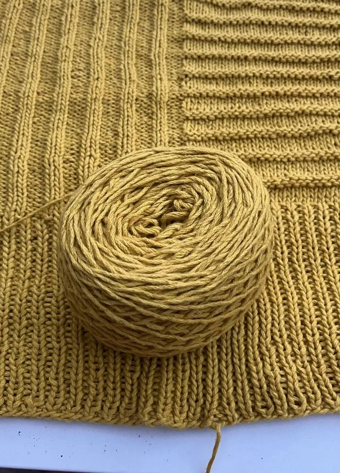 A ball of mustard yellow yarn sits atop a partially knit fabric with visible ribbing patterns, conveying a warm and cozy crafting vibe.