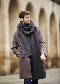 A man walks confidently wearing a long brown coat and a thick grey scarf in front of an arched stone building. The mood is stylish and serene.