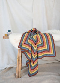 Colorful crochet blanket drapes over a wooden stool against a neutral fabric backdrop. Vertically stacked wooden blocks add a playful touch. Cozy and inviting.