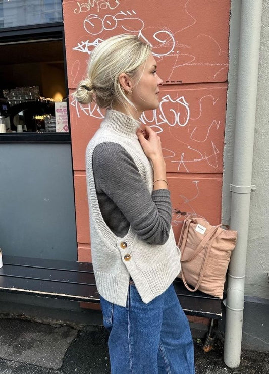 Woman in a grey knitted vest stands against a graffiti-covered wall, wearing jeans and holding a beige bag. Casual and artistic vibe.