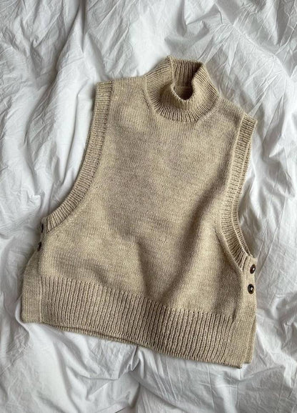 A beige knitted sleeveless turtleneck sweater vest lies on a crumpled white sheet, featuring side buttons and a cozy, minimalist aesthetic.