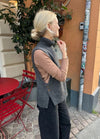 A woman with blond hair in a bun stands outside near a café, wearing a grey sleeveless sweater over a beige top. Red chairs and a potted plant are nearby.