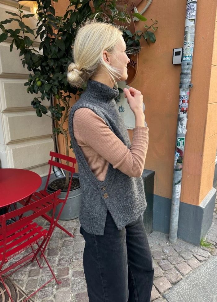A woman with blond hair in a bun stands outside near a café, wearing a grey sleeveless sweater over a beige top. Red chairs and a potted plant are nearby.