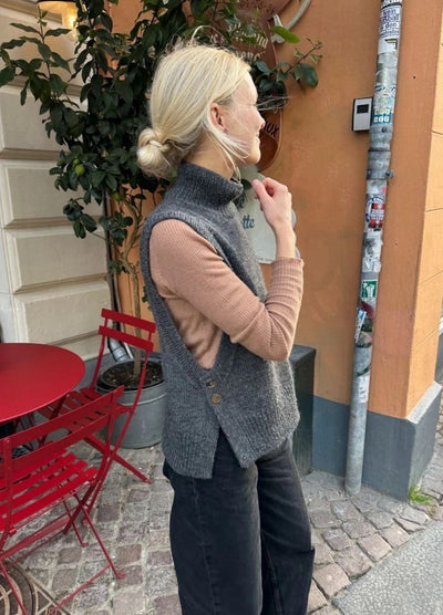 A woman with blond hair in a bun stands outside near a café, wearing a grey sleeveless sweater over a beige top. Red chairs and a potted plant are nearby.