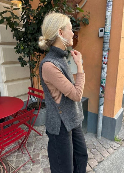 A woman with blond hair in a bun stands outside near a café, wearing a grey sleeveless sweater over a beige top. Red chairs and a potted plant are nearby.