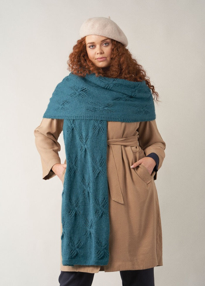 A woman with long curly auburn hair wears a camel trench coat layered with a large teal knitted wrap scarf decorated with subtle star-like stitch patterns. She also wears a light beige beret and stands with her hands in her pockets against a neutral background.
