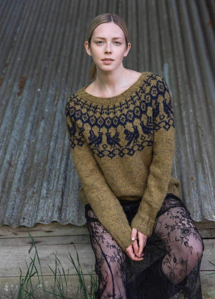 Maailman Synty Sweater: olive green knit with dark blue geometric and animal motifs around the yoke, worn with a sheer floral skirt.