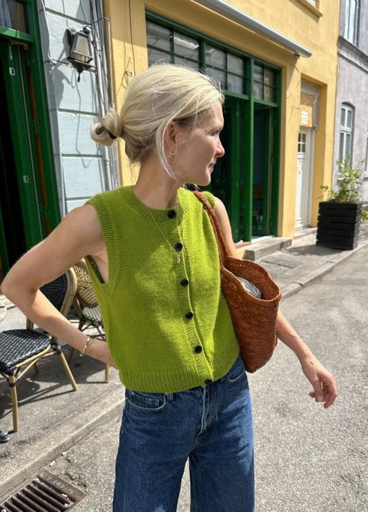 A woman in a bright green sleeveless top and jeans stands on a sunlit street, carrying a woven brown bag. The mood is casual and relaxed.