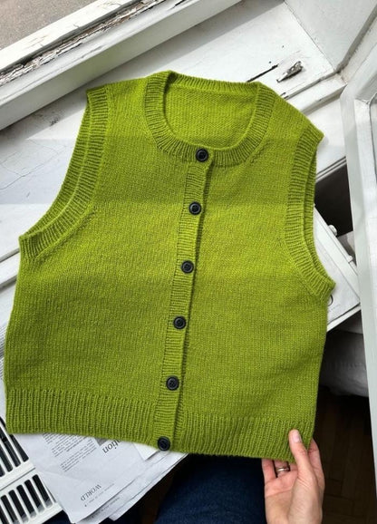 A bright green, sleeveless knit vest with black buttons is displayed on a white windowsill. A hand gently holds the bottom edge, conveying a sense of admiration.