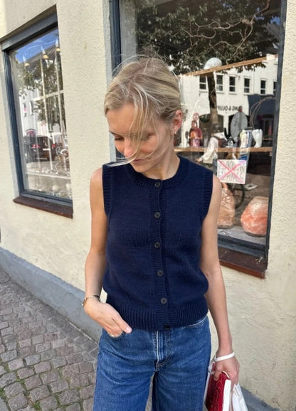 A woman in a navy sleeveless top and jeans stands on a cobblestone street, looking down. She is in front of a shop window displaying various items.