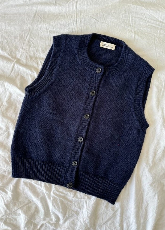 Navy blue knitted vest with buttons, laid on a crumpled white fabric background. The vest has a classic, sleeveless design, exuding a cozy, casual vibe.