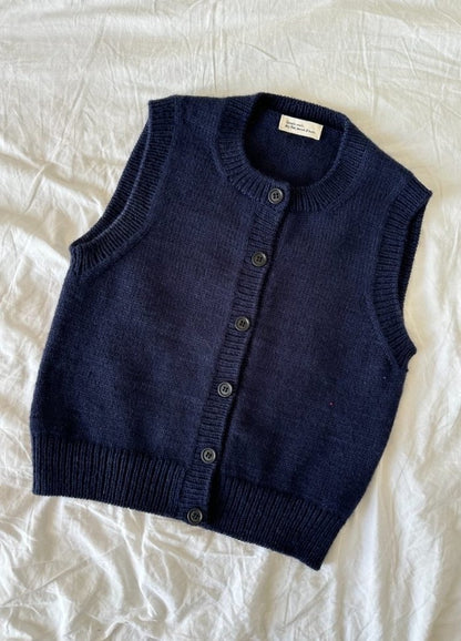 Navy blue knitted vest with buttons, laid on a crumpled white fabric background. The vest has a classic, sleeveless design, exuding a cozy, casual vibe.