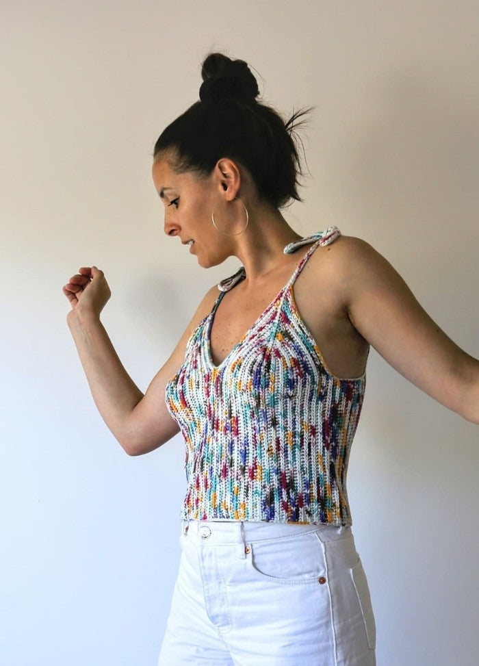 A woman poses confidently against a plain background, wearing a colourful, crochet tank top and white pants. Her hair is in a bun, and she has hoop earrings.