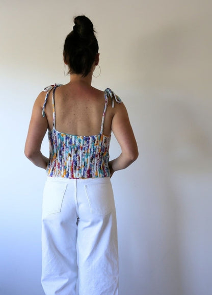 A woman with a bun stands facing a plain wall, wearing a colourful crochet tank top and white pants. Her upright posture gives a casual, relaxed vibe.