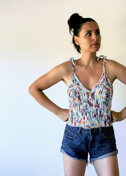Woman in a colourful, crochet tank top and denim shorts, stands confidently with hands on hips against a plain white background. She looks to the side.
