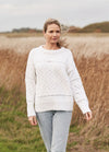 A woman walks through a grassy field, wearing a white knit sweater and light jeans. The wind blows her hair softly, creating a serene and peaceful mood.