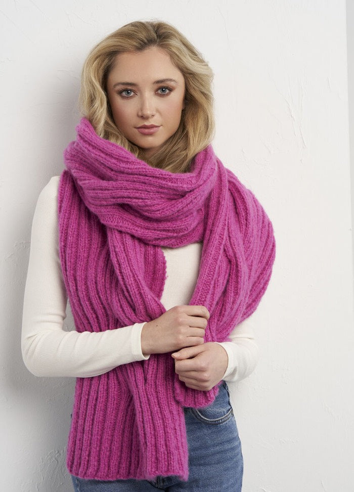 A woman with long blonde hair wears a thick, textured pink scarf over a white sweater. She stands in front of a white wall, looking serene and confident.