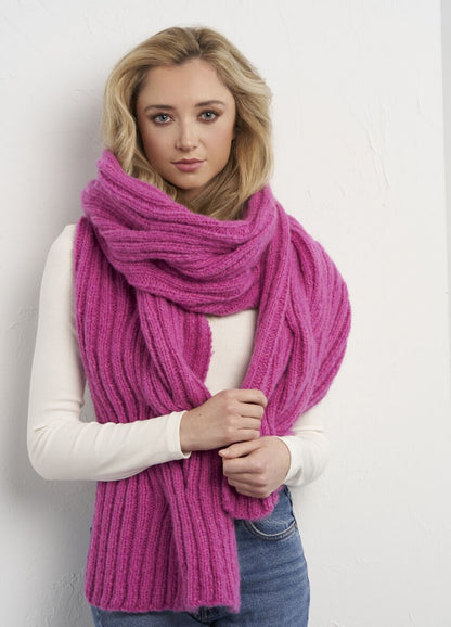 A woman with long blonde hair wears a thick, textured pink scarf over a white sweater. She stands in front of a white wall, looking serene and confident.