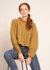 Person in mustard sweater and blue jeans sitting on a stool against a plain background.