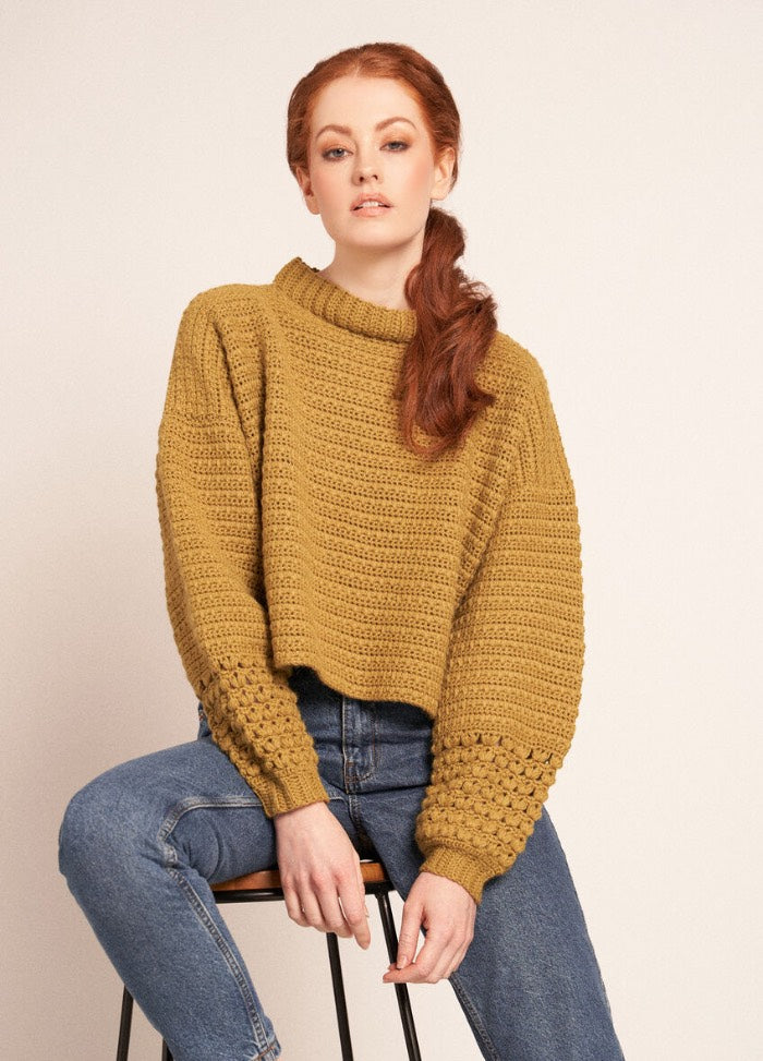 Person in mustard sweater and blue jeans sitting on a stool against a plain background.