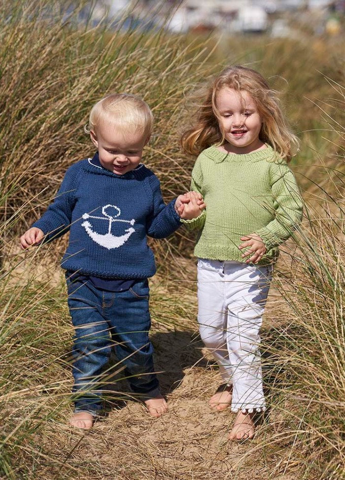Two young children walk hand-in-hand through tall grass on a sandy path. The child on the left wears a blue anchor sweater, while the one on the right wears a green sweater and white pants. They appear joyful and carefree.
