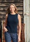 Woman in a navy sleeveless top and jeans stands confidently outdoors. She smiles softly, with a rustic wooden wall as the backdrop, creating a relaxed, casual vibe.