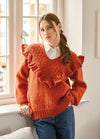 Young woman wearing an orange ruffled sweater and jeans leans against a windowsill. She gazes forward with a calm expression in a softly lit room.